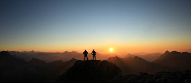 dois homens chegando ao cume desfrutando da liberdade e olhando para as montanhas ao pôr do sol. - sunset sun mountain sunrise - fotografias e filmes do acervo