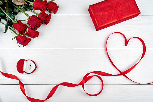 Red ribbon shaped as heart, bouquet of roses, red gift box and golden ring on white background, copy space. Valentines day, Womens Day. Love concept. Top view, flat lay