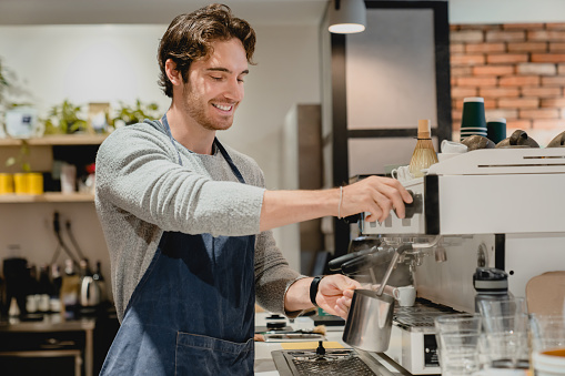 Cheerful young handsome barista making coffee on coffee machine in small cafeteria