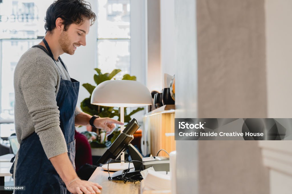 Side view portrait of young good-looking caucasian barista working on the cash point in cafeteria Cafe Stock Photo