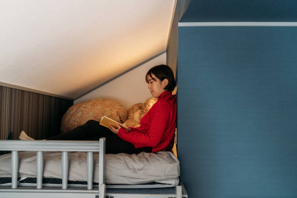 Young woman reading book at home at night A young woman is sitting on her bed comfortably and reading a book in a small loft apartment at home at night. japan small apartment stock pictures, royalty-free photos & images