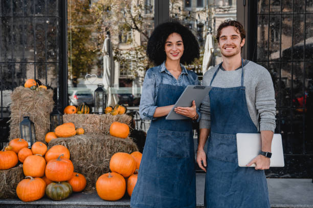 happy young coffee shop owners standing outdoor near their cafe with laptop and decorating pumpkins preparing for halloween on background. - holiday autumn season halloween imagens e fotografias de stock