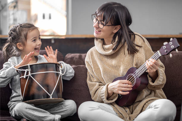 Young woman with her daughter have fun playing musical instruments at home. Cheerful mother and daughter are engaged in musical creativity, playing the drum and ukulele at home on the couch. Quality time together. drum percussion instrument photos stock pictures, royalty-free photos & images