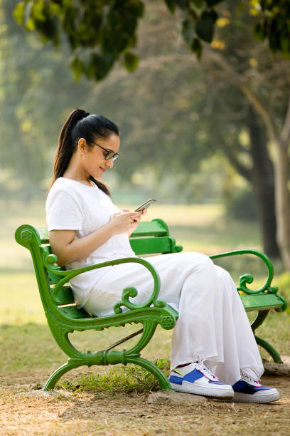 woman using mobile phone at park - using phone garden bench imagens e fotografias de stock