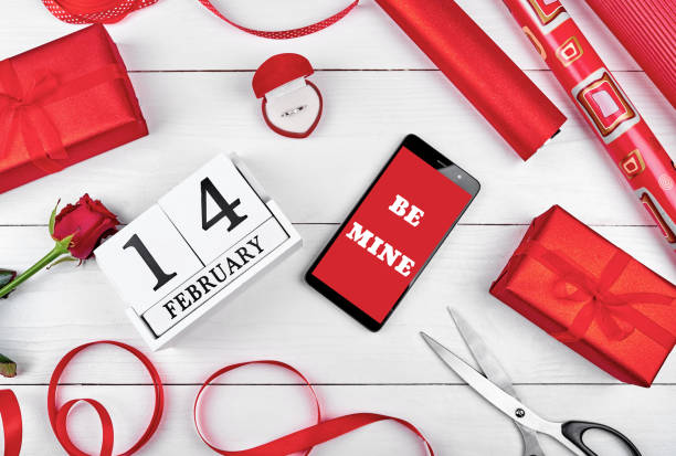 istock Gift wrapping background. Rolls of red paper, ribbon, red rose, giamond ring, gift boxes, mobile cellphone with sign Be mine and wood calendar february 14, free space. Flat lay, top view 1292409095