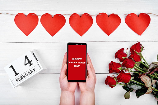 Girl holding mobile phone with sign Happy Valentines Day in hands, garland of red paper hearts, bouquet of roses and february 14 wooden calendar, copy space. Greeting card mockup. Top view, flat lay