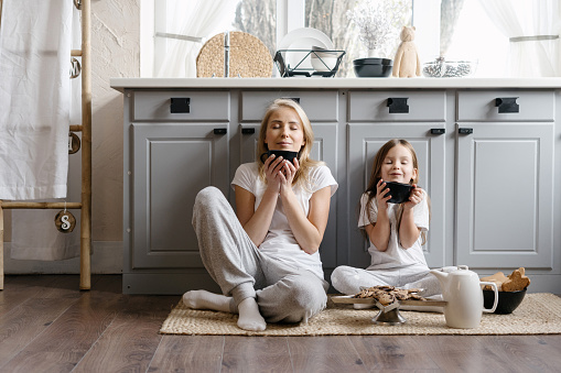 Dreamy mother sitting on kitchen floor together with her lovely daughter, enjoying morning coffee, drinking beverage and spending weekend in cozy apartment, resting at home