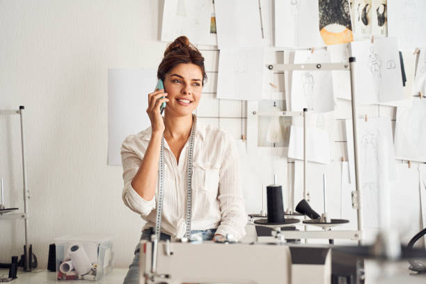 Designer standing in own dressmaking atelier Fashion entrepreneur. Happy brunette clothing designer standing in own dressmaking atelier and talking on the mobile phone. Copy Space woman tailor stock pictures, royalty-free photos & images