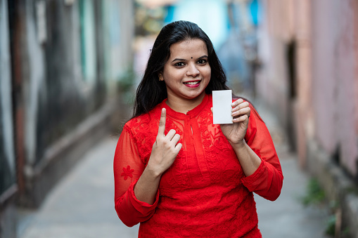 Outdoor image of an Indian woman showing her finer marked with ink and her voter id card after casting vote in Election.