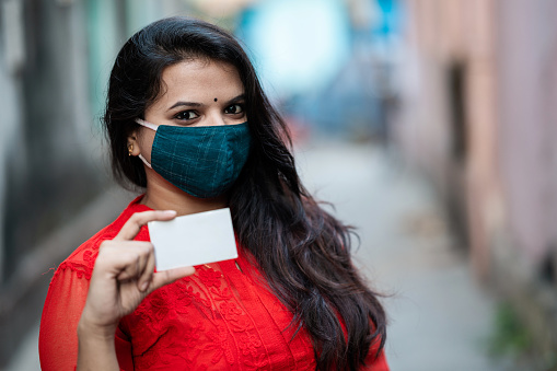 Young woman wearing face mask and showing business card. She is pointing on white card with other hand.