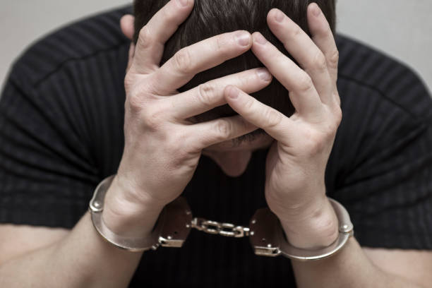 hombre esposado cubriéndose la cara con las manos. detenido y esposado criminal con esposas en las manos cubrir su cabeza y arrepentimiento en la estación de policía. - esposa fotografías e imágenes de stock