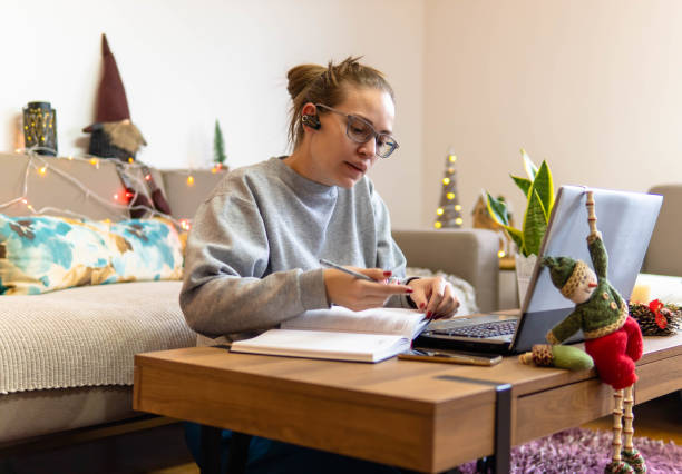 istock Remote work, office at home, work from home. Christmas time. A young woman uses a headset for an online talks, she sits on the floor with a laptop. Christmas tree and Santa. Coffee table. Earbuds 1292474162