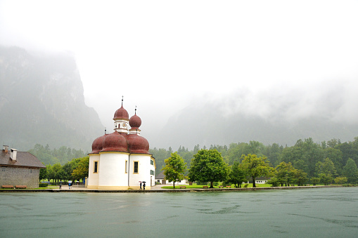 St. Bartholomew's church on Konigssee.  View from the water against the background of the mountains. Cloudy rainy day, fog, muddy dark water in the lake. Bavaria, Germany.