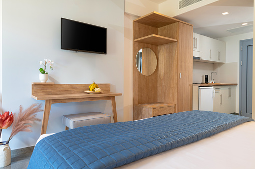 Modern style interior of small studio apartment. Hotel room with white kitchen, blue bedroom, pine wooden wardrobe, flat TV in single space