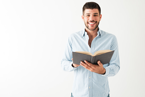 Hispanic young man wearing a shirt, smiling at the camera and holding an open book in front of a white background