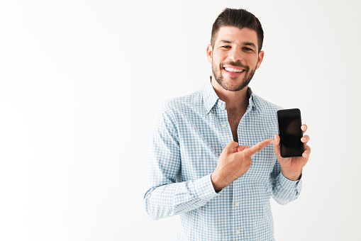 Portrait of a happy hispanic man in his 20s holding his smartphone and pointing to the screen