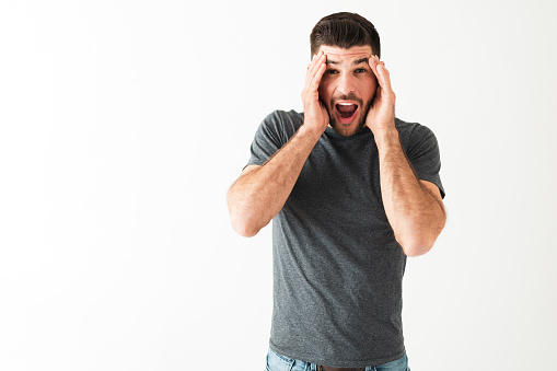 Hispanic man in his 20s looking with a shock and surprised expression. Young guy with his hands in the side of the face in front of a white background
