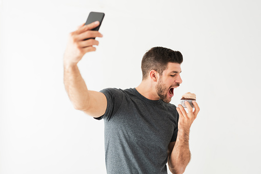 Attractive young man taking a selfie with his smartphone while eating a chocolate cupcake in front of a white background