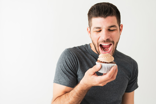 Good-looking hispanic man in his 20s taking a bite from a chocolate cupcake with frosting in front of a white background