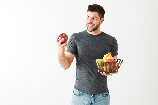 Hispanic young man is smiling and playing with an apple. Attractive man holding a basket of fresh fruits, isolated against a white background