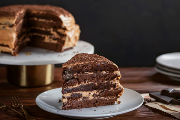 una fetta di torta al cioccolato brownie fatta in casa su un piatto bianco. la torta a strati è ricoperta di glassa al burro con latte condensato e gocce di cioccolato. piatti bianchi e forchette decorano il tavolo. - coffee cake foto e immagini stock