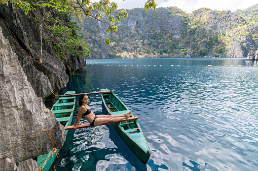 An athletic and beautiful mixed race woman traveling solo in a remote and beautiful location sits on a boat after taking a relaxing swim in a tropical lake surrounded by rocky cliffs.