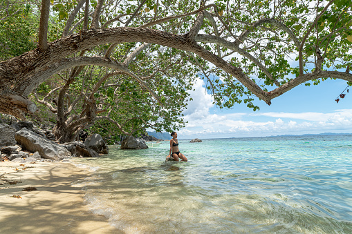 A beautiful mixed race young adult woman wearing a black bikini sits on a rock and dangles her feet in the water while relaxing at a remote tropical beach. The woman is traveling solo in the Philippines.