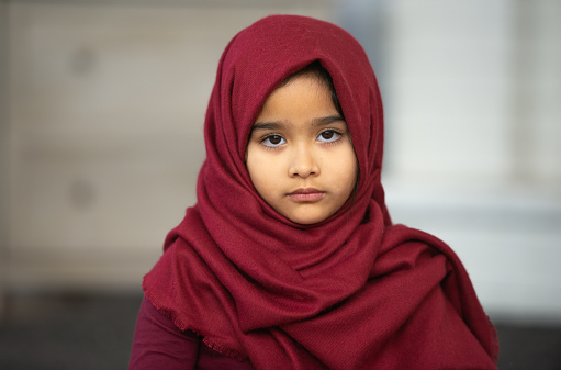 A serious looking toddler muslim girl looking at the camera. She is wearing a red hijab.