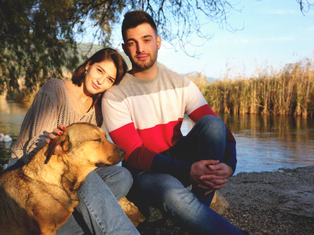 Young couple relaxing on river bank with visitor dog
