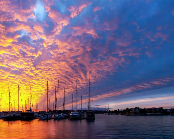A beautiful inspirational Gold Orange coloured cirrus cloud sunset seascape over silhouetted yachts in sea water with bright water reflections.\nPhotograph location Mooloolaba Marina, Queensland, Australia.