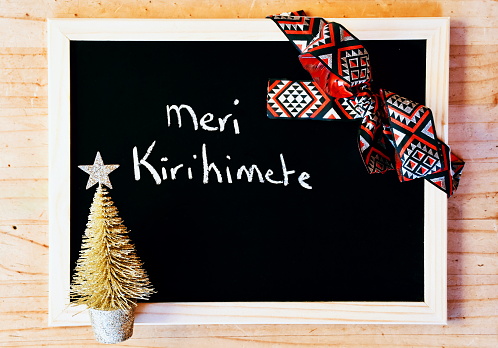 The words Meri Kirihimete (meaning Merry Christmas in Maori Te Reo) wrote on a Blackboard with Christmas Ribbon Bow & Christmas Tree Decoration.