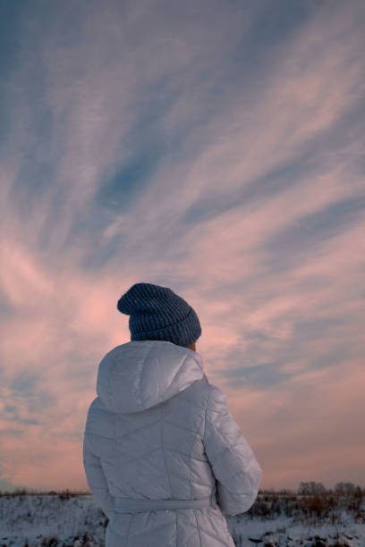 겨울, 필드, 눈, 저녁. 아름다운 일몰 하늘, 파란색과 분홍색 톤. 전경에는 흰색 재킷을 입은 소녀가 있습니다. 뒤에서 볼 수 있습니다. - russian ethnicity cold relaxation nature 뉴스 사진 이미지