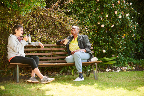Laughing senior friends sitting together on a park bench Two senior female friends talking and laughing together over coffees while social distancing on a park bench bench stock pictures, royalty-free photos & images