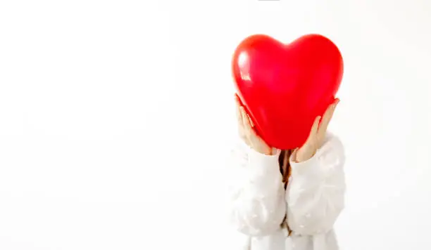 unrecognizable woman holding heart shaped ballon in front of his head, against white background in white sweater. Happy valentines day. love and relationships concept . 2021 Trend, banner