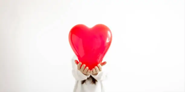 unrecognizable woman holding heart shaped ballon in front of his head, against white background in white sweater. Happy valentines day. love and relationships concept