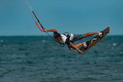 Carefree man having fun while jumping high-up with kiteboard during summer day at sea.