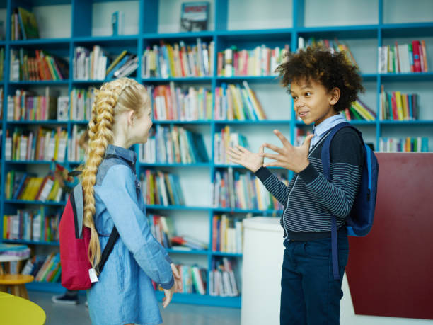 istock Two excited elementary students, mixed race curly-haired boy and blonde girl with braids, standing face-to-face near shelves in school library and talking 1292548513