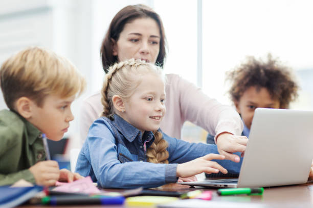 istock Female teacher explaining computer science to group of diverse elementary students looking at laptop screen sitting at desk in classroom 1292548524