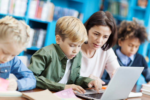 istock Side view of elementary student doing task on laptop computer helped by female teacher, his defocused classmates writing in notebooks sitting at desk in school classroom nearby 1292548530