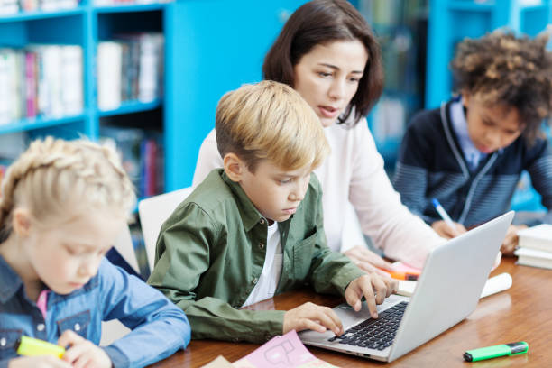 istock Elementary school boy doing task on laptop helped by female teacher, his classmates writing in notebooks sitting at desk in classroom nearby 1292548532
