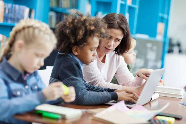 istock Side view selective focus of female teacher explaining task on laptop screen to mixed race elementary student, his classmates studying sitting at desk in school classroom nearby 1292548538