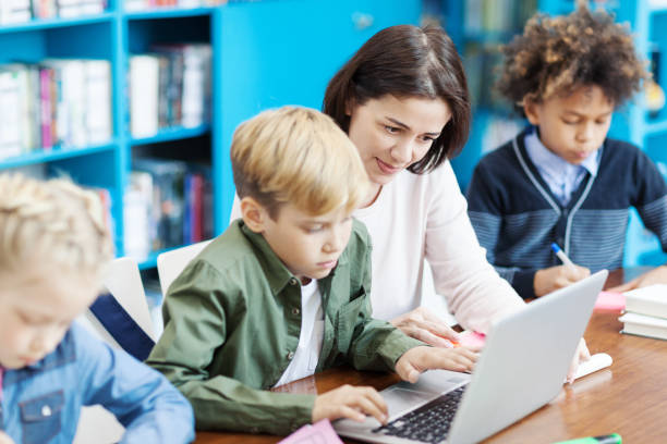 istock Side view of elementary student doing task on laptop helped by female teacher, his classmates writing in notebooks sitting at desk in school classroom nearby 1292548540