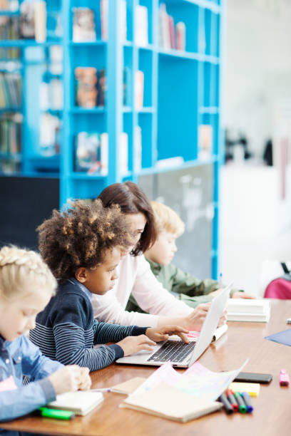 istock Side view of female teacher explaining task on laptop computer screen to mixed race elementary student, his classmates studying sitting at desk nearby in school classroom 1292548544
