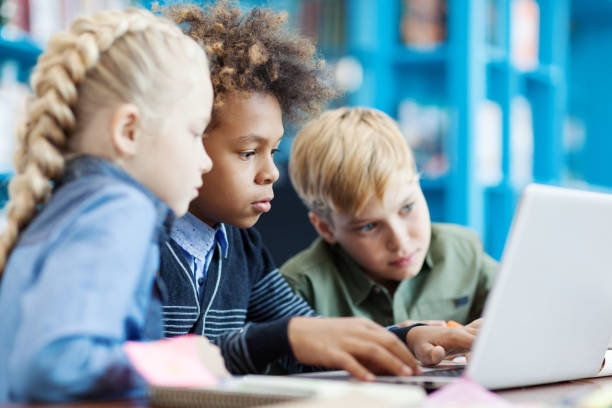 istock Side view of three curious diverse students using laptop sitting at desk in school library. Mixed  race schoolboy and his friends browsing through information on Internet 1292548547