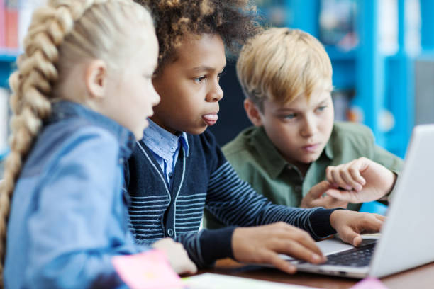 istock Side view of three curious diverse students with laptop sitting at desk in elementary school classroom. Mixed  race schoolboy and his friends searching for information on Internet 1292548548