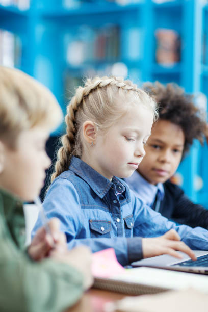 istock Group of three elementary students sitting at desk in school library and studying; focus on little blonde girl typing on laptop surrounded by diverse male friends 1292548553