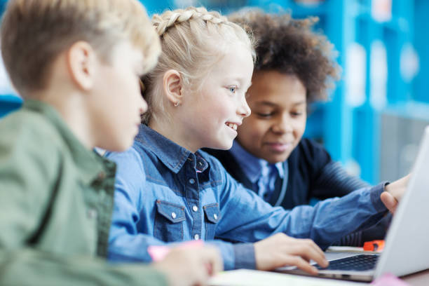 istock Group of three elementary students sitting at desk in classroom and studying or playing on laptop; focus on smiling blonde girl pointing at screen 1292548555