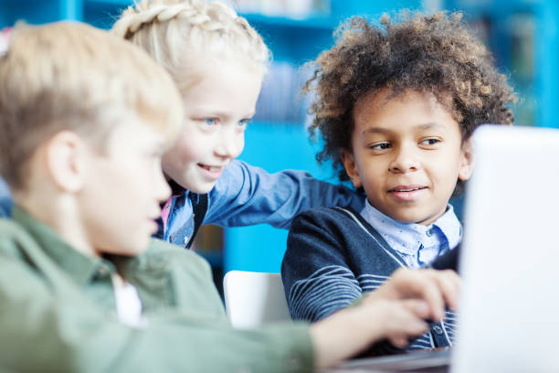 istock Side view of three curious little school students, two boys and girl, browsing Internet on laptop in school library. Focus on mixed race boy explaining something to friends 1292548585
