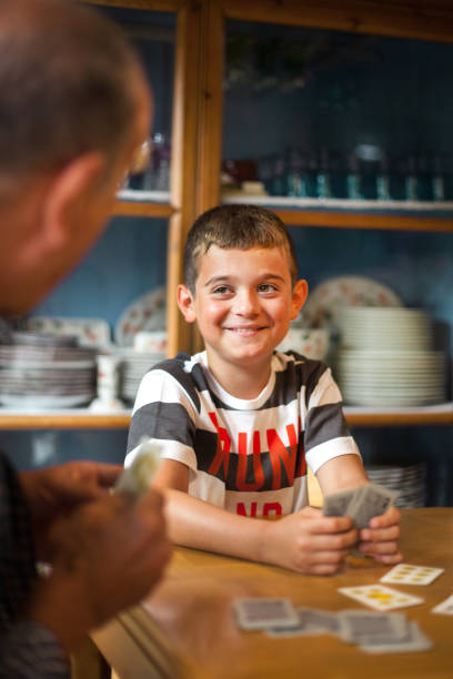 8 years-old boy sitting around a table, playing cards with his family. Kid with a big smile looking at his grandfather while playing a board game. It's daytime.