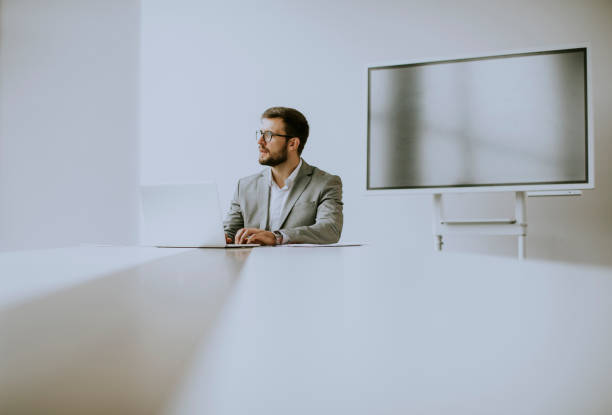 istock Young man working on laptop in bright office with big screen behind him 1292554940